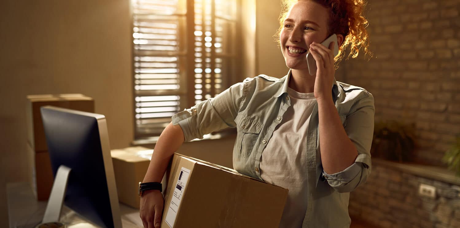 A woman holds a box while speaking on the phone, appearing engaged in a conversation.